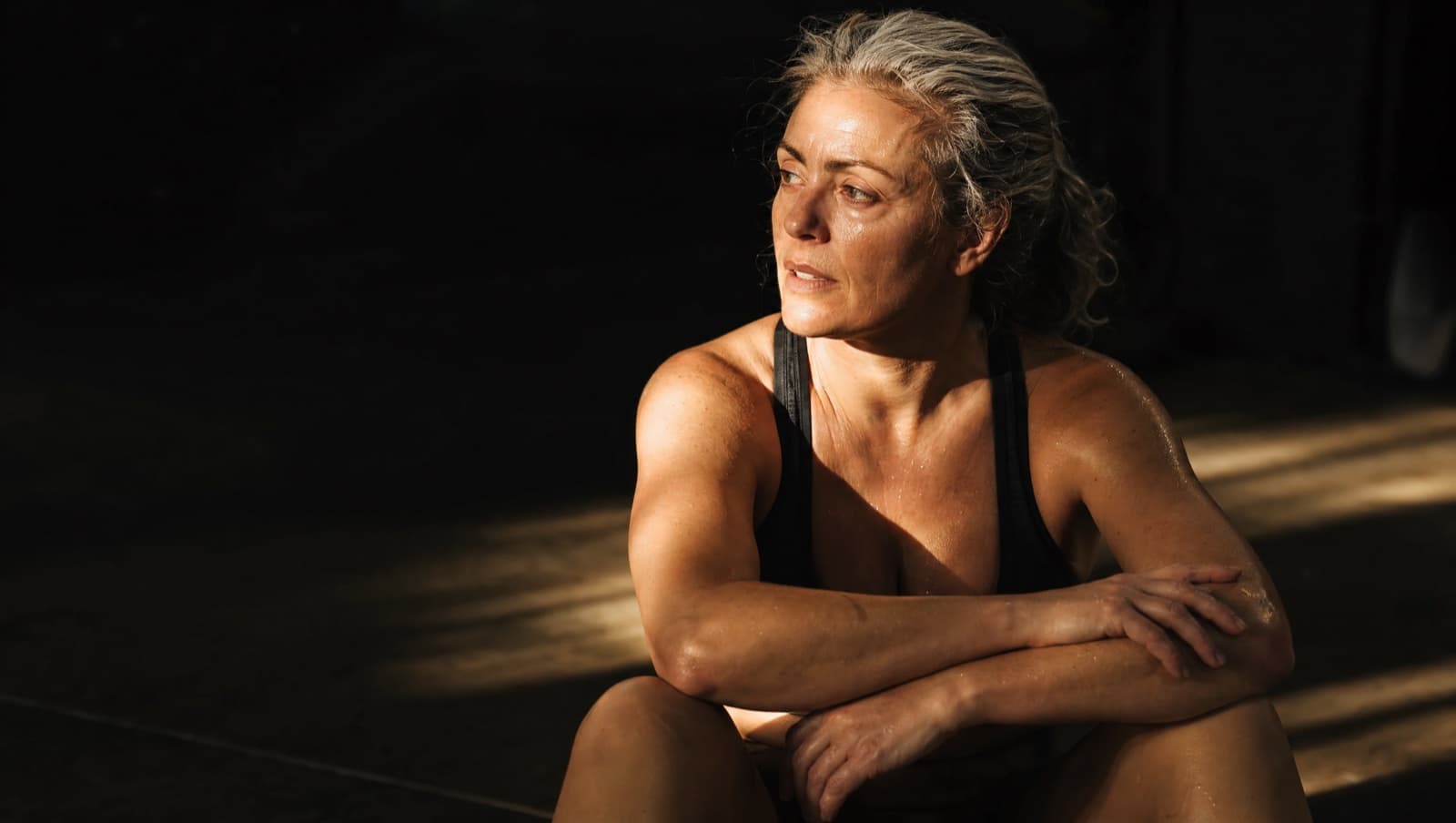 A woman with grey-streaked hair sits on a gym floor after training, forearms on her knees, catching her breath, rim-lit from the side against a dark background.