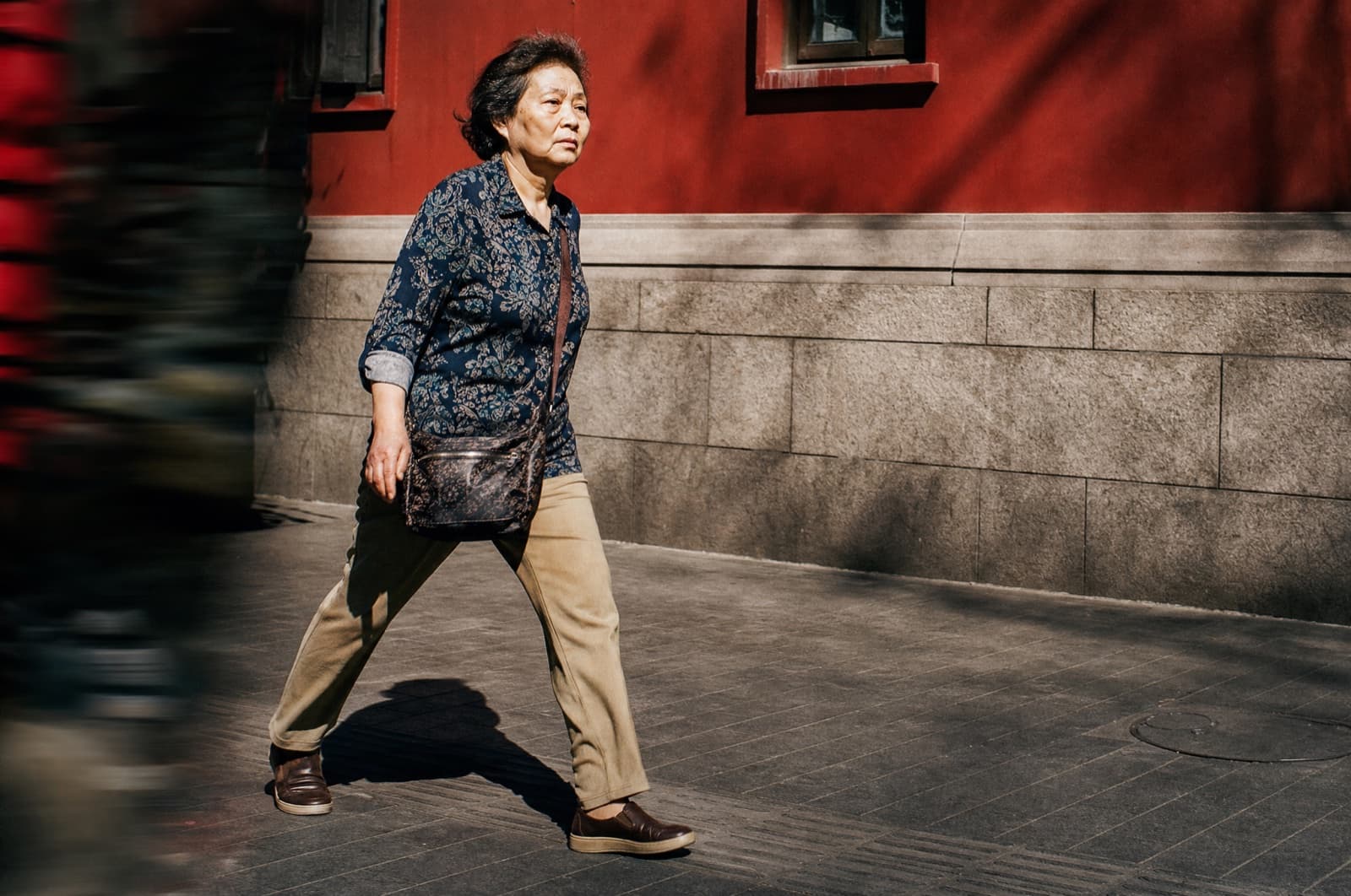 A woman in her sixties walks a city sidewalk at midday with a brisk, purposeful stride, hard sunlight throwing a crisp shadow across the pavement.