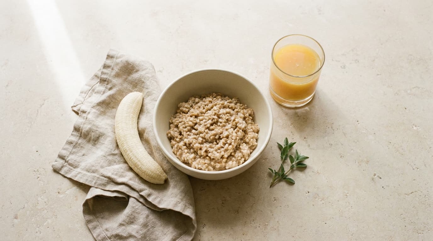 Overhead still life in warm window light: a bowl of steel-cut oatmeal, a peeled banana on a linen napkin, and a glass of orange juice.