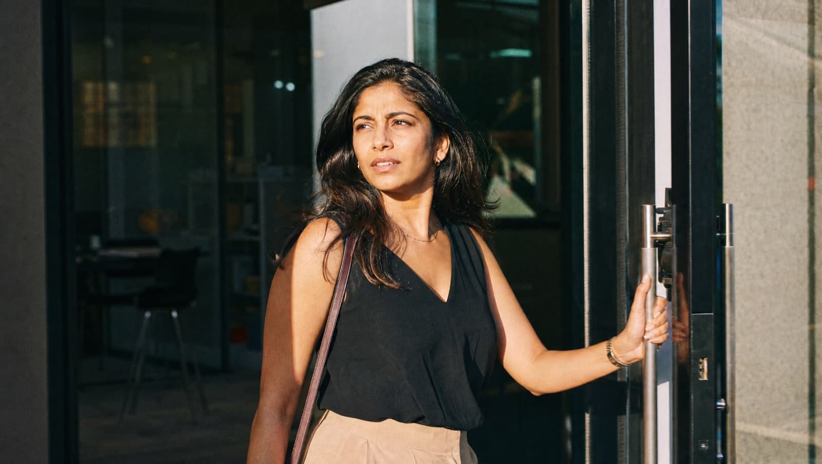 A woman pushes open a glass office door into bright midday sunlight, caught mid-step, backlight flaring warm around her.