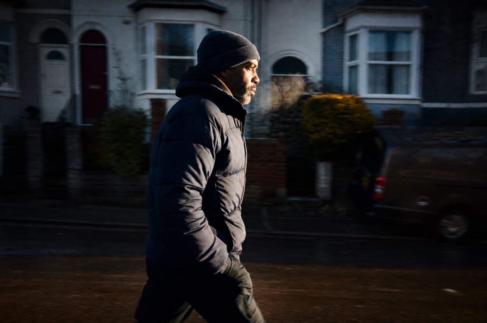 A man in his forties runs mid-stride along an ordinary residential street in cold early light, breath visible, the background blurred by motion.