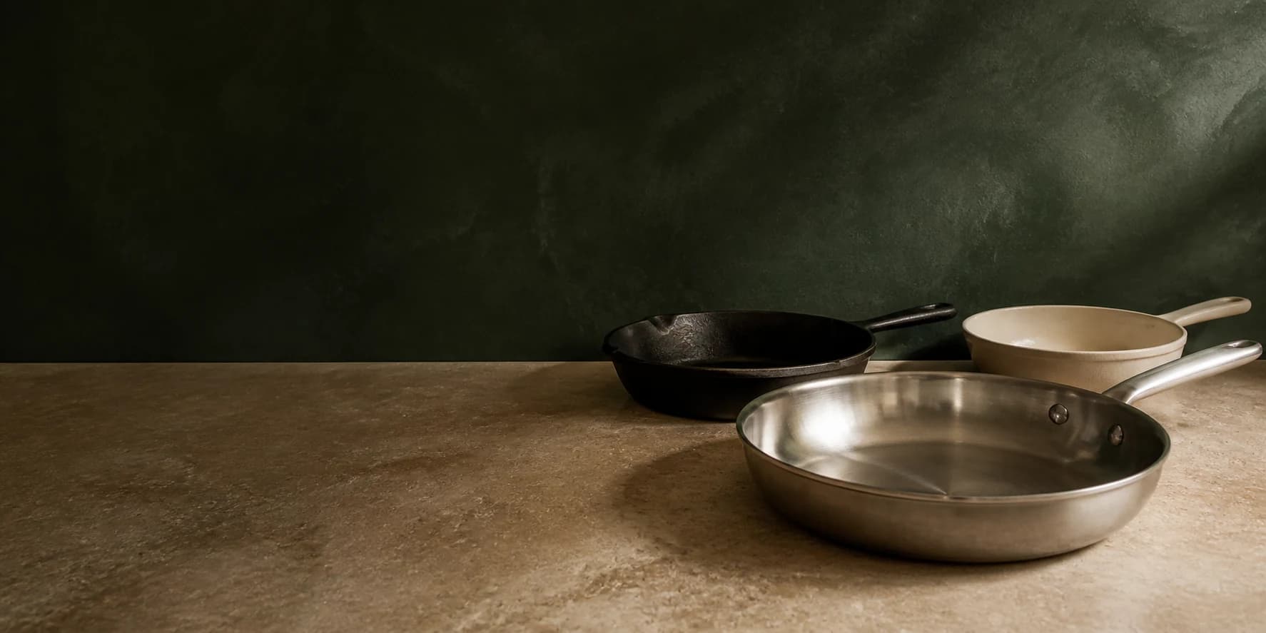 Stainless steel, cast iron, and cream ceramic skillets on stone against a deep green wall.