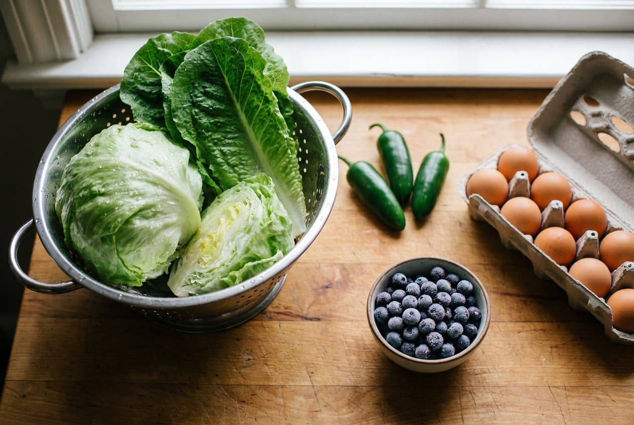 Overhead view of a kitchen counter with rinsed lettuce in a colander, whole jalapeños, a bowl of frozen blueberries, and brown eggs in an open carton in soft window light.
